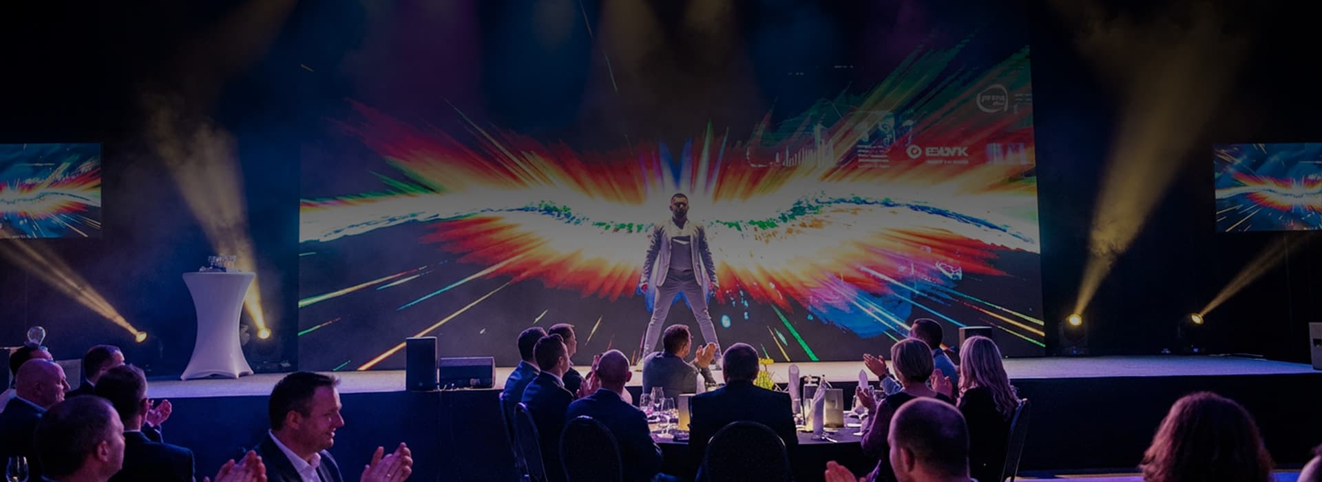 Ein Mensch steht auf einer Bühne vor einem bunten, leuchtenden Hintergrund, mit applaudierenden Zuschauern im Vordergrund.
